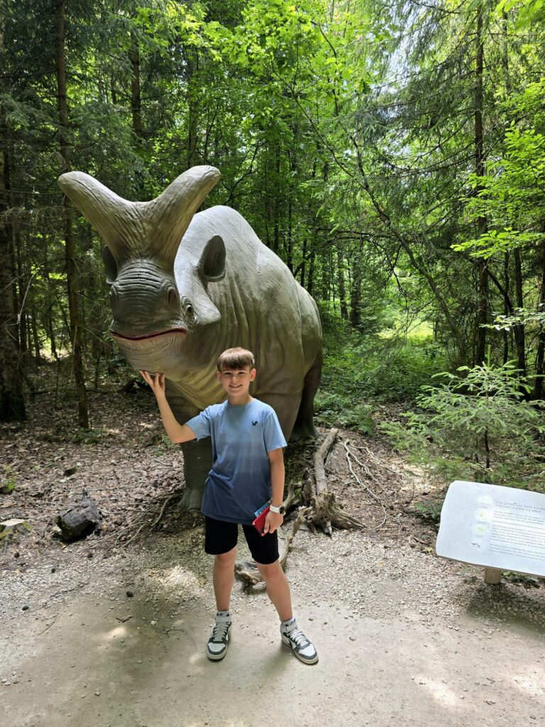 Kind posiert vor einer großen Dino-Skulptur im Wald des Dinosauriermuseums Altmühltal. Beliebtes Familienausflugsziel in Bayern.