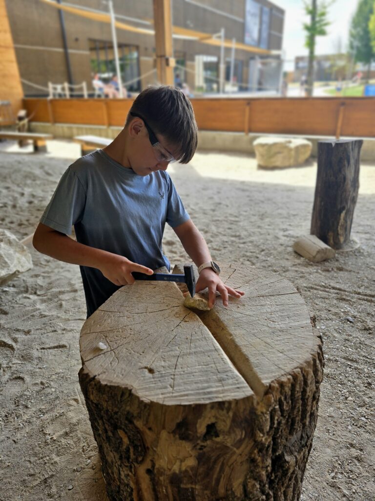 Kind arbeitet mit Hammer und Meißel an einem Holzstamm im Dinosauriermuseum Altmühltal. Interaktive Mitmachstation für Kinder.