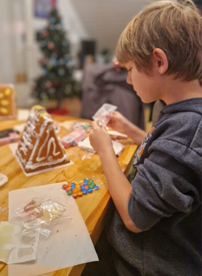 Ein Kind sitzt konzentriert am Tisch und dekoriert sein Lebkuchenhaus mit bunten Süßigkeiten, während im Hintergrund schon der Weihnachtsbaum leuchtet.