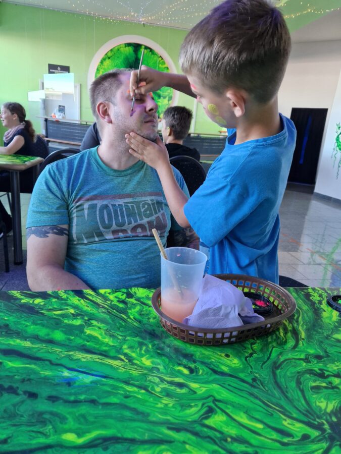 Vater sitzt an einem Tisch in der Schwarzlichtfabrik Nürnberg, während sein Sohn ihm mit Neonfarbe das Gesicht bemalt.