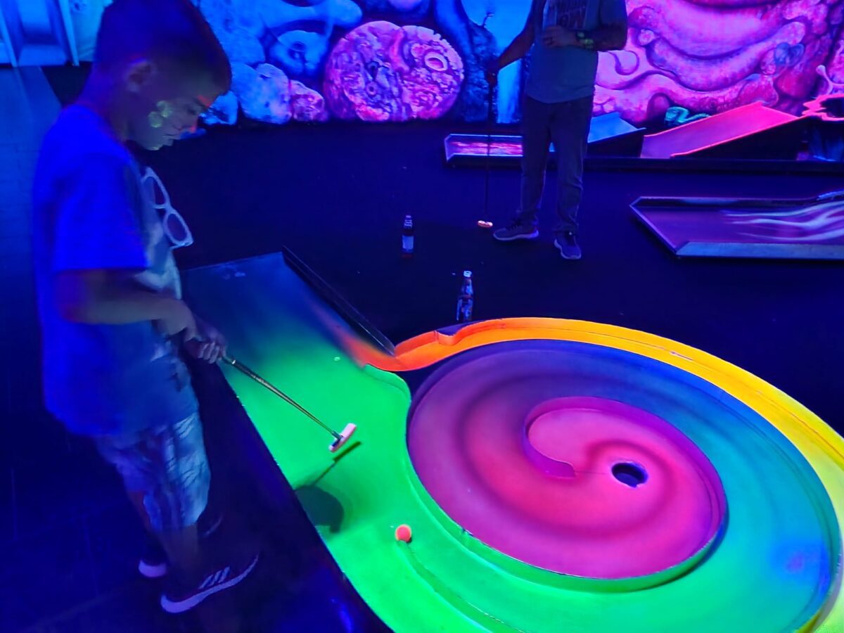 Kind spielt auf einer leuchtenden Schwarzlicht-Minigolfbahn in bunten Neonfarben.