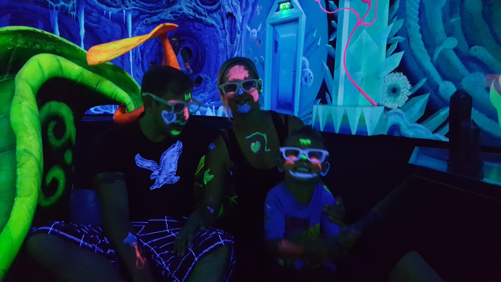 Familie sitzt im schwarzlichtbeleuchteten Minigolfbereich, trägt Neonbemalung im Gesicht und weiße 3D-Brillen.