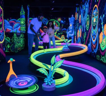 Familie spielt 3D-Schwarzlichtminigolf auf einer bunt leuchtenden Neonbahn mit starken UV-Farben und fantasievollen Wandmotiven.