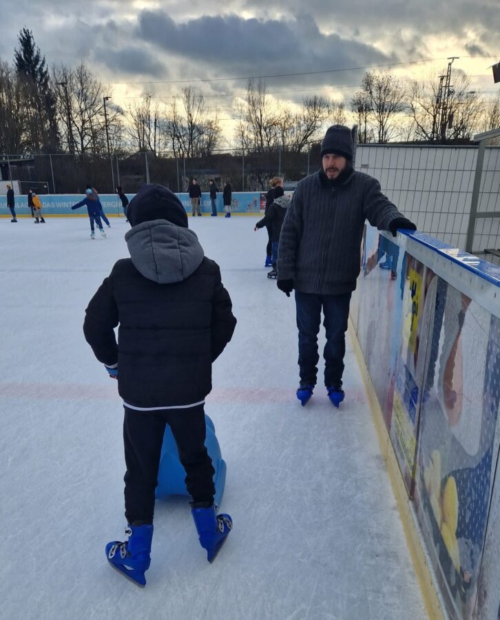 Kind steht mit Eislaufhilfe an der Bande der Kunsteislaufanlage in Neumarkt, im Hintergrund weitere Schlittschuhläufer.