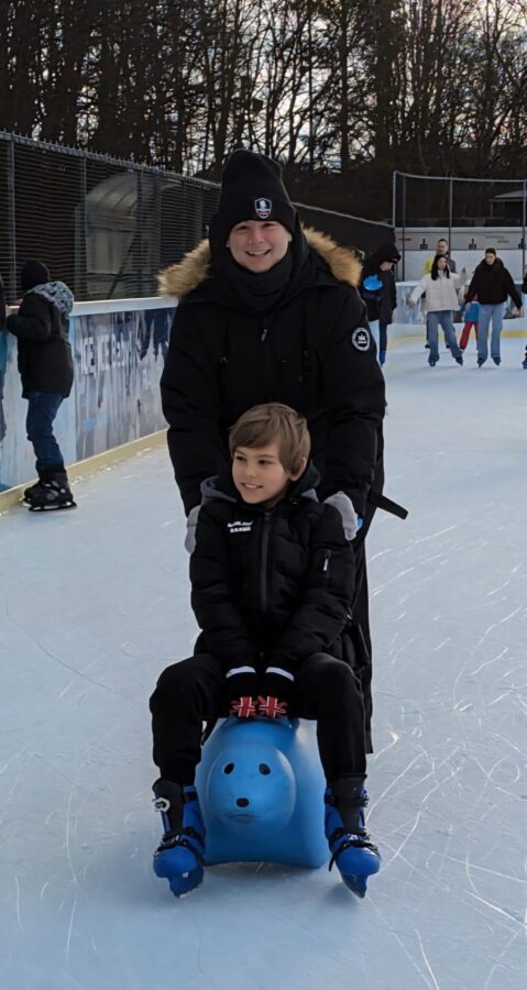 Erwachsener schiebt ein Kind lachend auf einem blauen Eislauf-Seehund über die Eisfläche in Neumarkt.